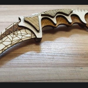 Könnte beinhalten: Ein Holzmesser mit einem Spinnennetz-Design. Das Messer ist aus hellbraunem Holz gefertigt und hat ein weißes Spinnennetz-Muster auf der Klinge. Der Griff ist ebenfalls aus Holz und hat ein braunes und weißes Muster.