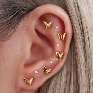 May include: Close-up of an ear adorned with multiple gold butterfly earrings and small diamond studs. The butterfly earrings are in various sizes and positions, creating a layered effect. The word "OPVARA" is visible in the image.