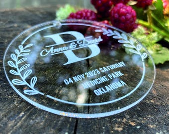 May include: A clear round acrylic ornament with the text "Anna & David" and "04 NOV 2023 SATURDAY MEDICINE PARK OKLAHOMA" in white lettering.
