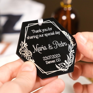 May include: Black hexagonal tag with white floral and geometric designs. Text reads "Thank you for sharing our special day" and "Maria & Pedro" with a date and location. A white ribbon is attached.