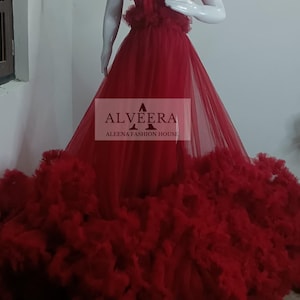 May include: A strapless, floor-length red gown with a layered, ruffled skirt and bodice. The dress is displayed on a mannequin. The text "ALVEERA ALEENA FASHION HOUSE" is visible on the image.