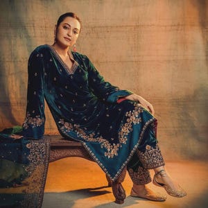 May include: A woman wearing a teal blue velvet kurta with gold embroidery and matching trousers. She is sitting on a wooden bench and wearing gold flats.