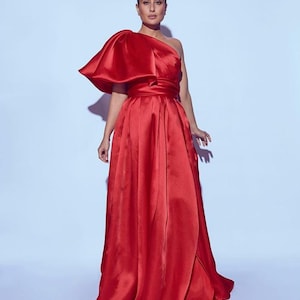 May include: A vibrant red, one-shoulder gown with a voluminous, draped bodice and a flowing skirt. The dress features a cinched waist and a dramatic, oversized shoulder detail. The fabric has a satin sheen, reflecting light. The gown is floor-length.