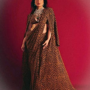 Puede incluir: Una mujer con un conjunto de estampado de leopardo marrón y negro, que incluye una capa y una falda larga. También lleva un collar grande y pendientes. El fondo es de color rojo liso.