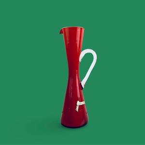 May include: A red glass pitcher with a white handle. The pitcher has a narrow neck and a wide base.