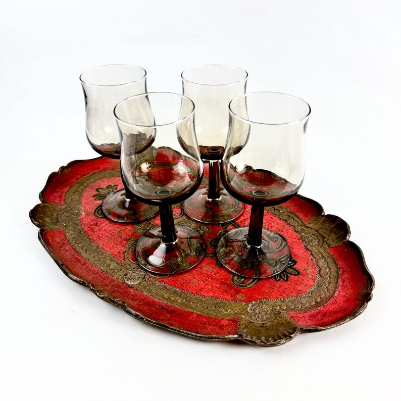 May include: Four clear glass wine glasses with dark brown stems sit on a red and gold decorative tray. The tray has a scalloped edge and intricate designs.