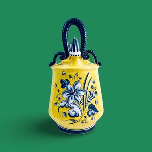 Könnte beinhalten: Eine gelbe Keramikkanne mit einem blauen und weißen Blumendesign und einem schwarzen Henkel. Die Kanne hat einen Ausguss und einen Deckel.
