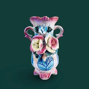 Puede incluir: Un jarrón decorativo de cerámica con un diseño floral. El jarrón presenta una combinación de colores rosa claro y azul con rosas y hojas esculpidas. Tiene dos asas y un borde festoneado, con una altura aproximada de 20 cm.