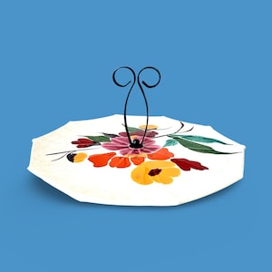 Puede incluir: Un soporte para tartas de cerámica blanca con un diseño floral con flores rojas, amarillas, naranjas y rosas. El soporte tiene un asa de metal negro.