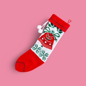 May include: A festive Christmas stocking with a red cuff and toe, and a white body. The body features a red bell design with green accents and snowflakes. A white loop and two white pom-poms are attached to the top.