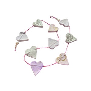 Könnte beinhalten: Eine Girlande aus herzförmigen Ornamenten in verschiedenen Weiß-, Rosa- und Lavendeltönen, auf einer rosa Schnur aufgereiht. Einige Herzen haben strukturierte Designs oder goldene Akzente. Die Girlande ist kreisförmig vor weißem Hintergrund angeordnet.