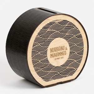Puede incluir: Un banco de dinero de madera marrón oscuro con una ranura en la parte superior. La parte delantera presenta un diseño circular con un patrón de ondas y los nombres "NURBANU & MUHAMMED" y la fecha "03 MART 2023" grabados en un círculo.