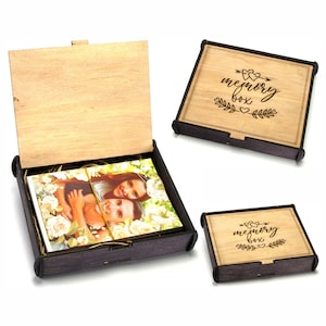 Può includere: Scatola dei ricordi in legno con coperchio a cerniera, contenente fotografie. La scatola è marrone scuro con un coperchio marrone chiaro. Il coperchio è inciso con le parole "memory box" ed elementi decorativi. La scatola è aperta, rivelando foto all'interno.