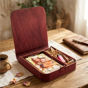 May include: A deep red stained wooden box with an upright hinged lid. The interior is divided into two compartments, one containing a rectangular photo print of a couple surrounded by white flowers, and the other holding dried raffia and a small pink object.