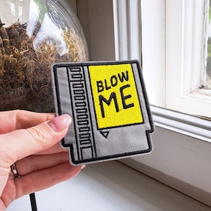 Könnte beinhalten: Ein gestickter grauer und weißer Patch in Form einer Videospielkassette mit einem gelben Rechteck auf der Vorderseite, auf dem in schwarzer Schrift "BLOW ME" steht.