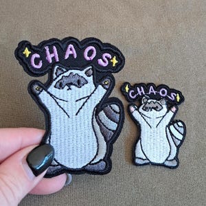 Op de afbeelding: Twee geborduurde patches met een lichtgrijze wasbeer met donkergrijze oogvlekken en een gestreepte staart. Boven de wasbeer staat het woord CHAOS in lichtpaarse letters op een zwarte achtergrond, geaccentueerd door twee kleine gele sterren.