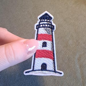 Könnte beinhalten: Gestickter Aufnäher eines Leuchtturms mit rot-weißen Streifen, einem blauen Oberteil und einem weißen Rand. Der Aufnäher hat ein maritimes Thema und ist für Kleidung oder Accessoires geeignet.