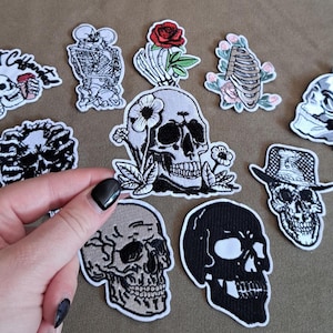 Può includere: Una collezione di toppe ricamate con vari disegni di teschi, tra cui uno con una rosa rossa, uno con girasoli, uno con un cilindro e uno con il testo Dead Inside But Caffeinated, tutti caratterizzati da dettagliate cuciture in bianco e nero.