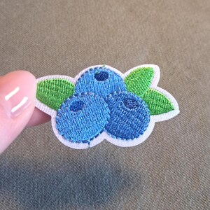 Könnte beinhalten: Gestickter Aufnäher mit drei blauen Blaubeeren und grünen Blättern. Der Aufnäher hat einen weißen Rand und ist zum Aufbügeln oder Aufnähen auf Stoff vorgesehen. Ideal zum Aufpeppen von Kleidung oder Accessoires.