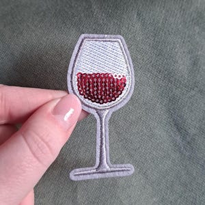 Può includere: Toppa ricamata a forma di calice di vino. Il bicchiere è delineato in grigio con una parte superiore ricamata bianca e "vino" con paillettes rosse che riempie la metà inferiore. La toppa è tenuta su uno sfondo verde.