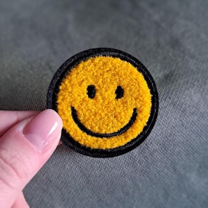 Könnte beinhalten: Ein runder, gelber Smiley-Aufnäher mit schwarzem Rand. Das Smiley-Gesicht hat zwei schwarze Augen und einen geschwungenen schwarzen Mund. Der Aufnäher besteht aus einem strukturierten Material.