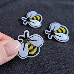 May include: Three embroidered bee patches. Each patch features a white body, yellow and black striped abdomen, and black antennae. The patches are on a dark gray fabric.