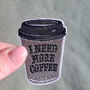 May include: Embroidered patch in the shape of a coffee cup with a black lid. The cup is gray with the words "I NEED MORE COFFEE" embroidered in black. The patch is on a green background.