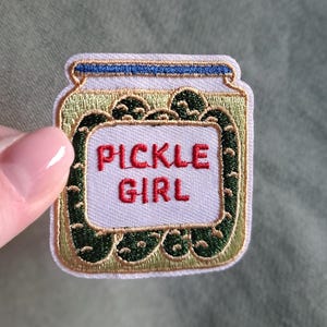 Könnte beinhalten: Gestickter Aufnäher in Form eines Einmachglases mit den Worten "PICKLE GIRL" in Rot. Das Glas hat einen grünen und goldenen Rand mit einem blauen Deckel. Der Aufnäher befindet sich auf einem grünen Hintergrund.