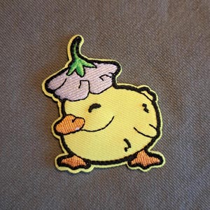 Op de afbeelding: Een gele geborduurde patch met een lachend, rond karakter met oranje voeten en een bijpassende hand. Het karakter draagt een paarse bloemenhoed met een groene stengel. De patch heeft een zwarte omtrek.