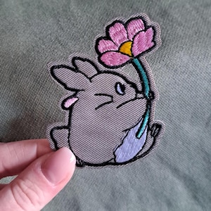 Könnte beinhalten: Gestickter Aufnäher mit einem grauen Cartoon-Hasen, der eine rosa Blume mit gelbem Zentrum hält. Der Hase hat schwarze Umrisse und einen hellvioletten Bauch. Der Aufnäher befindet sich auf einem grünen Hintergrund.