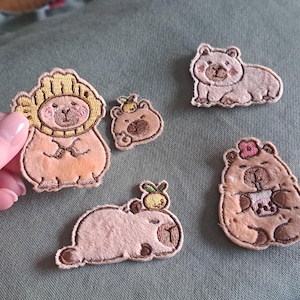 Può includere: Una collezione di toppe ricamate raffiguranti capibara dei cartoni animati in varie pose. Le toppe sono nei toni del marrone, beige e pesca, alcune con accessori come una criniera di leone o un fiore. Le toppe sono piccole e dettagliate.