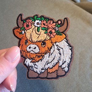 Könnte beinhalten: Gestickter Aufnäher mit einer Cartoon-Hochlandkuh mit Blumenkranz. Der Aufnäher hat einen braunen Rand, das Fell der Kuh ist in Orange-, Weiß- und Beigetönen gehalten. Die Blumen im Kranz sind rosa, orange und weiß.