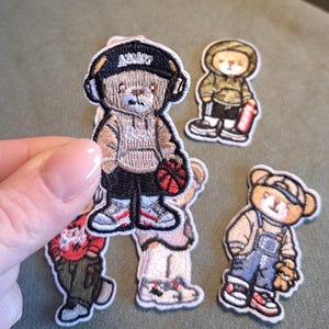 Könnte beinhalten: Gestickte Aufnäher mit Cartoon-Teddybären in verschiedenen Outfits. Ein Bär trägt einen Kapuzenpullover, eine Kappe und Kopfhörer und hält einen Basketball. Andere Bären sind in Streetwear mit Turnschuhen gekleidet. Die Aufnäher haben detaillierte Stickereien.
