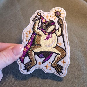 May include: Embroidered patch featuring a whimsical frog wizard. The frog is depicted in a cream-colored robe and a purple cape with gold stars. It holds a staff with a sunburst design. The patch has a white border with gold star accents.