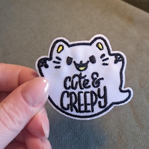 May include: A white embroidered patch shaped like a ghost with cat ears, black stitched outlines, and yellow accents. The patch features the black embroidered text "cute & CREEPY" in the center, with a small black mouth and whiskers.