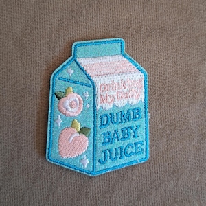 Könnte beinhalten: Ein gestickter Aufnäher in Form eines hellblauen Milchkartons. Er zeigt zwei rosa Pfirsich-Illustrationen, kleine weiße Sternakzente sowie den Text "Drink My Daily" und "DUMB BABY JUICE" in blauem und rosa Garn.