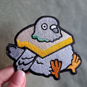 May include: Embroidered patch depicting a cartoon pigeon. The patch features grey and black feathers, orange feet, and a yellow and tan collar. The design is whimsical and suitable for clothing or accessories.