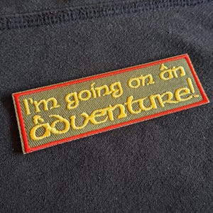 May include: Embroidered patch with the text "I'm going on an adventure!" in yellow thread on an olive green background, framed by a red border. The patch is rectangular and sewn onto a dark fabric.