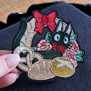 Könnte beinhalten: Gestickter Aufnäher mit einem schwarzen Katzendesign, umgeben von Blumen, einer roten Schleife und einer Brezel. Der Aufnäher hat einen grünen Rand und wird von einer Hand gehalten. Das Design ist farbenfroh und detailliert.