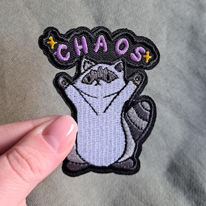 Könnte beinhalten: Gestickter Aufnäher mit einem Cartoon-Waschbären mit ausgestreckten Armen. Der Aufnäher hat das Wort "CHAOS" in lila Buchstaben über dem Waschbären, mit gelben Sternakzenten. Der Waschbär ist helllila, mit einer schwarzen Maske und einem Schwanz.