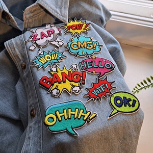 Könnte beinhalten: Eine Jeansjacke mit bunten gestickten Aufnähern in Form von Sprechblasen im Comic-Stil mit Wörtern wie "Zap!" "Pow!" "OMG!" "Hello!" "Bang!" "WTF!" "Ok!" und "Ohhh!"