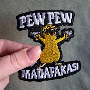 Könnte beinhalten: Gestickter Aufnäher mit den Worten "PEW PEW MADAF*KAS!" in weißem Faden auf schwarzem Hintergrund. Der Aufnäher zeigt einen gelben Cartoon-Vogel, der zwei Pistolen hält. Der Aufnäher ist rechteckig mit schwarzem Rand.