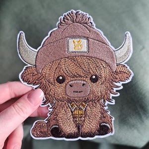 Op de afbeelding: Geborduurde patch met een cartoon Highland-koe met een bruine hoed met een pompon en een gouden embleem. De koe heeft grote ogen, gebogen hoorns en een gedetailleerde, getextureerde bruine vacht. De patch heeft een witte rand.