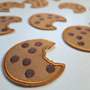 May include: A set of nine brown fabric patches shaped like chocolate chip cookies with a bite taken out of them. The patches have a brown fabric background with brown stitching around the edges and dark brown chocolate chips.