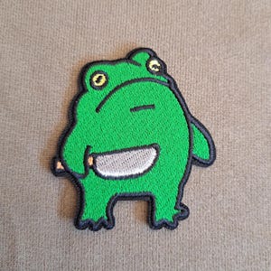 Könnte beinhalten: Ein gestickter Aufnäher, der einen leuchtend grünen Frosch mit gelben Augen und einer schwarzen Umrandung zeigt. Der Frosch ist stehend dargestellt und hält ein silberfarbenes Messer mit weißer Klinge in der rechten Hand vor einem einfarbigen Hintergrund.