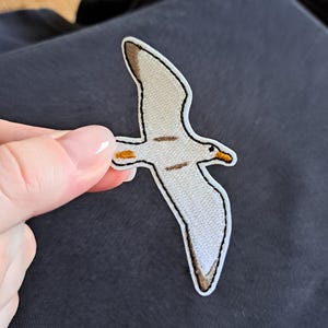 Op de afbeelding: Geborduurde patch van een vliegende zeemeeuw. De patch heeft een witte body met bruine en oranje accenten, omlijnd met zwarte stiksels. De zeemeeuw wordt tegen een donkerblauwe achtergrond gehouden.