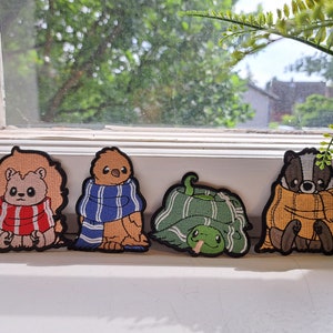 Può includere: Quattro toppe ricamate con personaggi animali che indossano sciarpe. I personaggi sono un leone, un uccello, una tartaruga e un tasso. Le toppe sono tutte di colori diversi e hanno uno stile fantasioso.