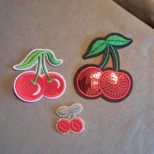 May include: Three embroidered cherry patches of different sizes. The largest is embellished with red sequins, the others have a smooth red finish. All have green leaves and stems, with white or gold trim.