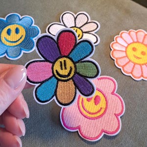 May include: Embroidered flower patches in various colors, including blue, white, pink, and a rainbow design. Each patch features a smiling face in the center, with a white border.