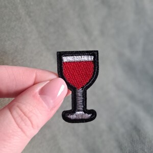 Può includere: Toppa ricamata raffigurante un bicchiere di vino rosso con un contorno nero. Il bicchiere è riempito di liquido rosso e presenta un bordo bianco nella parte superiore. La toppa è piccola e per abbigliamento o accessori.